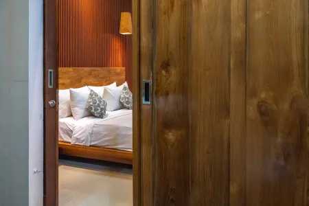 Cozy wooden bedroom interior through open slatted door, with king bed, white linens, gray pillows, and warm wall lamp.