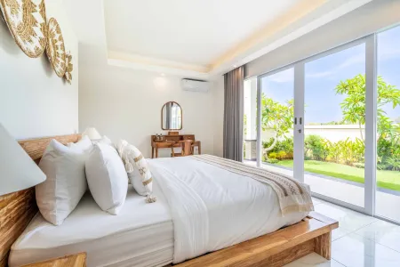 Spacious Balinese bedroom with wooden bed, white linens, vanity mirror, and sliding doors opening to tropical garden.
