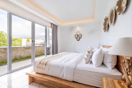 A serene Balinese-style bedroom featuring a wooden platform bed with crisp white linens, bedside lamp, and sliding glass doors opening to a lush tropical balcony view.