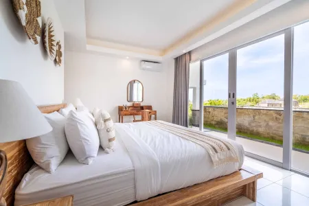 Spacious Balinese bedroom with wooden platform bed, white linens, vanity mirror, and floor-to-ceiling doors opening to tropical garden view.