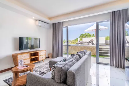Modern Bali villa living room with gray sofas, wooden accents, and sliding doors opening to an infinity pool deck with lounge chairs. - Bali Villas