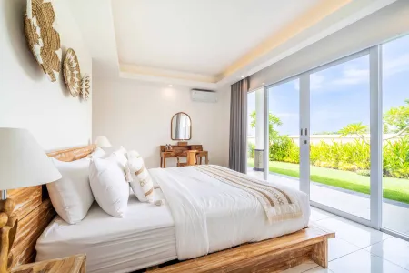 Serene tropical Bali bedroom with wooden platform bed, white linens, woven wall art, and sliding glass doors to lush garden view. - Bali Villas