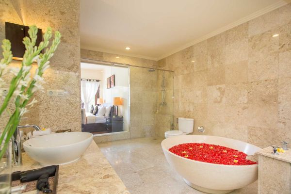 Luxurious Balinese-style bathroom with petal-filled bathtub, glass shower, double sinks, and bedroom view through sliding doors. - Bali Villas