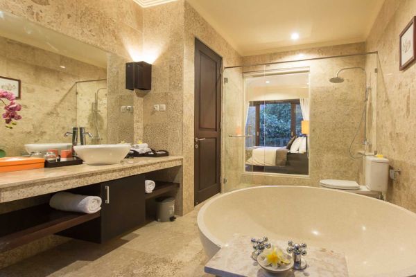 Luxurious Balinese bathroom with freestanding tub, stone vanity, orchids, and garden-view bedroom beyond glass doors.