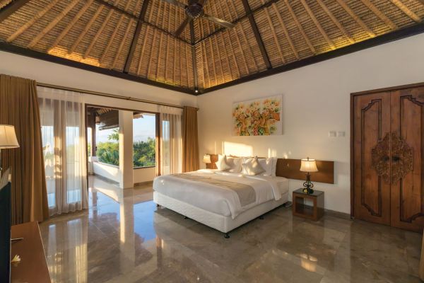 Luxurious Balinese villa bedroom with thatched roof, white king bed, carved wooden doors, and tropical balcony view.