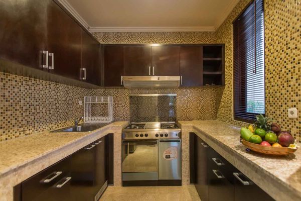 Modern U-shaped kitchen with dark wood cabinets, gold mosaic tile backsplash, granite counters, gas stove, and a bowl of fresh tropical fruits. - Bali Villas