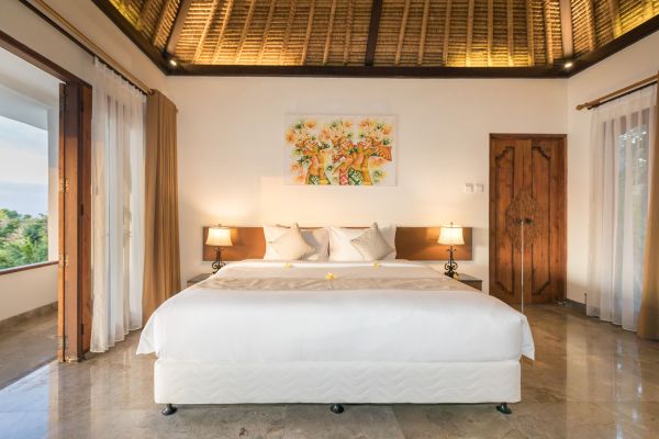 Luxurious Balinese villa bedroom featuring a king bed with frangipani accents, thatched ceiling, abstract wall art, and tropical views through sheer-curtained doors.