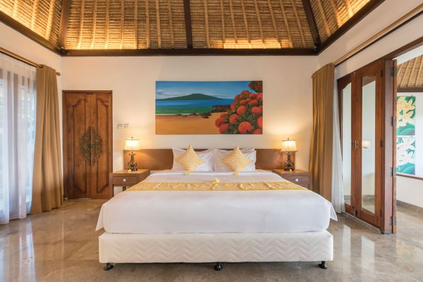 Luxurious Balinese bedroom with thatched ceiling, king bed topped with yellow frangipani, tropical beach painting, and sliding doors to balcony.