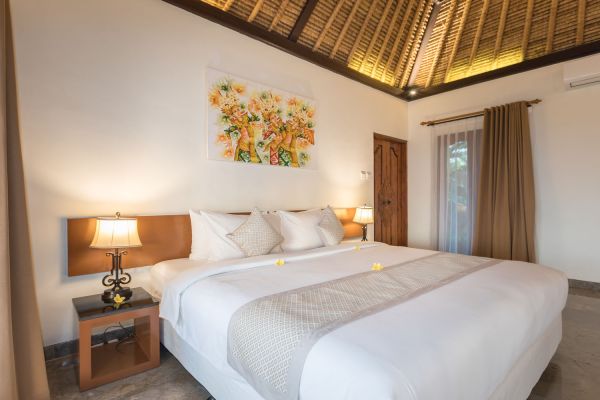 Spacious Balinese villa bedroom with king-size white bed, frangipani flowers, wooden lamps, floral wall art, and thatched ceiling.