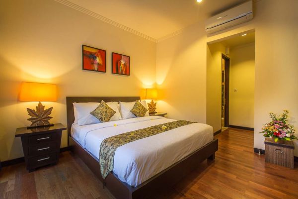 Cozy Balinese-style hotel bedroom with king-size bed, tropical lamps, abstract orange wall art, and potted flowers on hardwood floors.