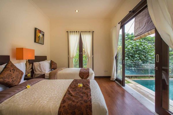 Elegant Balinese villa bedroom with plush bed, orange lamp, and sliding doors to infinity pool with tropical garden view.
