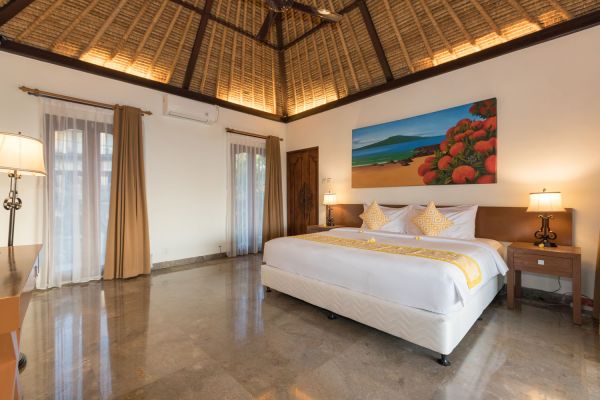 Spacious Balinese villa bedroom with king bed, thatched ceiling, tropical seascape painting, and open balcony doors.