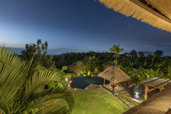 Twilight view from a thatched Balinese pavilion overlooking an illuminated infinity pool, lush tropical gardens, and distant ocean horizon at dusk. - Bali Villas