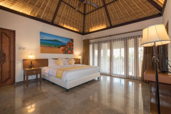 Luxurious Balinese villa bedroom with thatched ceiling, king bed, tropical wall art, and sliding glass doors to outdoor terrace.