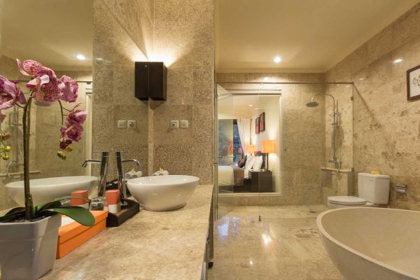 Luxurious hotel bathroom with beige stone tiles, double vanity, freestanding bathtub, orchids, and bedroom view.