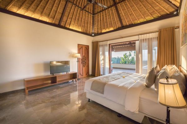 Spacious Balinese villa bedroom with king-sized bed, wooden furniture, TV, and open balcony overlooking tropical greenery.