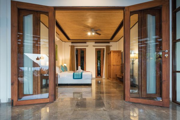 Spacious Balinese bedroom with open wooden doors to balcony, king bed, ceiling fan, and polished marble floors.