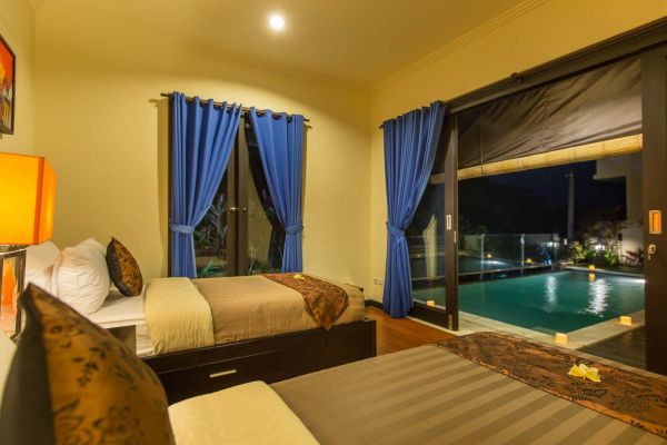 Cozy Bali villa bedroom with twin beds, blue curtains, and nighttime infinity pool view through sliding doors.