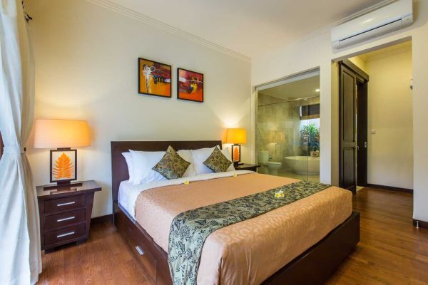 Spacious Balinese-style bedroom with wooden king bed, orange bedding, tropical lamps, abstract wall art, and glass-door en-suite bathroom.