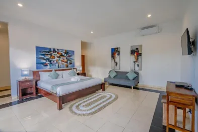 Spacious luxury bedroom in a tropical villa with king-size wooden bed, abstract wall art, gray sofa, mounted TV, and tiled flooring.