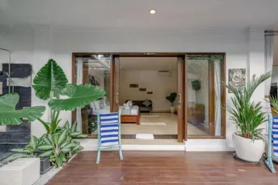 Luxury tropical villa bedroom with open sliding doors to a wooden deck, flanked by large potted plants and blue lounge chairs.