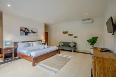 Spacious Balinese villa bedroom with wooden king bed, gray loveseat, potted plant, abstract art, and wall-mounted TV.