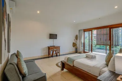 Spacious tropical bedroom with wooden bed, gray sofa, TV desk, and sliding wooden doors opening to lush greenery.