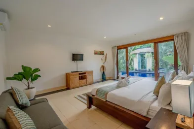 Serene Bali villa bedroom with king-sized wooden bed, tropical plants, TV stand, and sliding doors opening to an infinity pool.