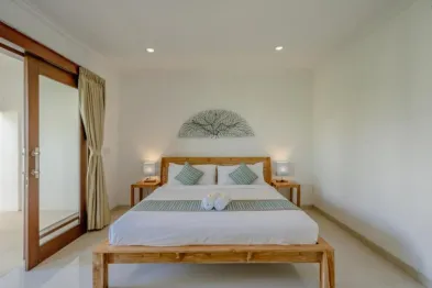 Cozy king bed with teal pillows and runner in a serene Bali-style bedroom with white walls and sliding wooden doors.