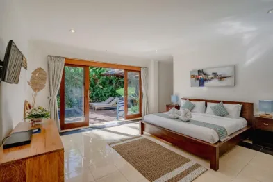 Spacious tropical bedroom with king-size wooden bed, open French doors to lush garden pool, and modern Bali-style decor.