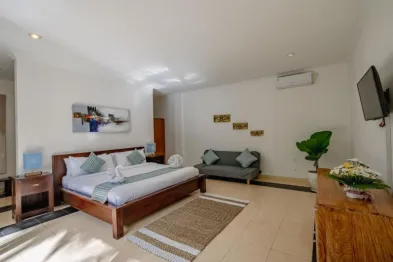 Spacious Balinese-style hotel bedroom with wooden king bed, gray loveseat, abstract wall art, potted plant, fresh flowers, and mounted TV in a bright tropical room.