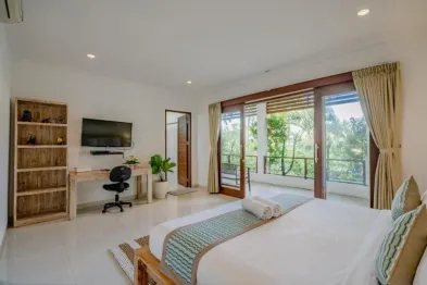 Spacious Balinese villa bedroom with king bed, wooden desk, and sliding doors opening to lush tropical balcony view.