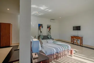 Bright, airy Balinese-style bedroom with king bed, wooden furniture, wall art, TV, and natural sunlight streaming in.