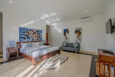 Spacious sunlit bedroom with wooden king bed, abstract wall art, gray sofa, desk, and tiled floor in a luxury villa.