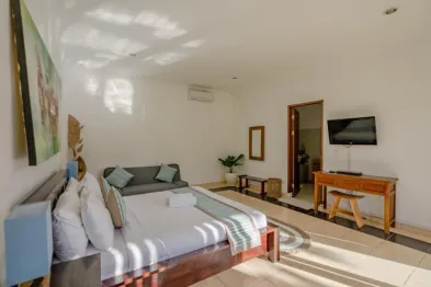 Spacious Balinese-style hotel bedroom with king bed, gray sofa, wooden desk, TV, and tropical decor.