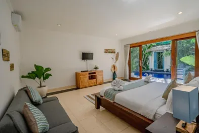 Spacious Bali villa bedroom with king bed, teal accents, sofa, TV, and sliding doors to tropical pool.
