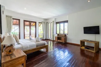 Spacious tropical bedroom with wooden king bed, white linens, large sliding glass doors to lush greenery, and polished hardwood floors.