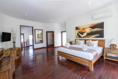 Spacious Balinese villa bedroom with wooden king bed, abstract winged figure wall art, hardwood floors, and tropical accents.