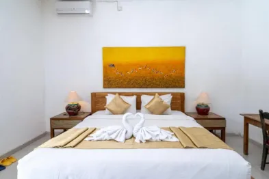 Cozy hotel bedroom with king bed adorned by white towel swans, golden bedding, wooden nightstands, and vibrant yellow abstract painting on white wall.