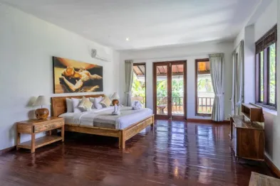 Spacious Balinese bedroom with wooden king bed, abstract wall art, open French doors to tropical balcony, and polished teak floors.