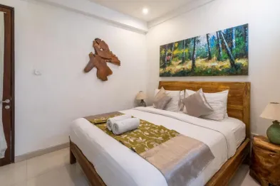 Cozy Bali villa bedroom with wooden king bed, white linens, forest painting, and carved wood decor.