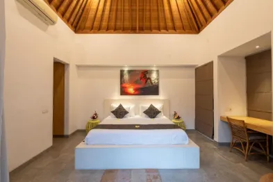 Spacious Balinese villa bedroom with high wooden ceiling, king bed with red pillows, abstract sunset wall art, and rattan desk.