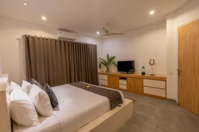 Modern minimalist bedroom with white king bed, wooden cabinets, TV, potted plant, and large curtained windows in a tropical villa.