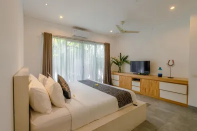 Spacious modern Bali bedroom with white king bed, wooden cabinets, flat-screen TV, potted plants, and floor-to-ceiling windows with sheer curtains overlooking lush greenery.