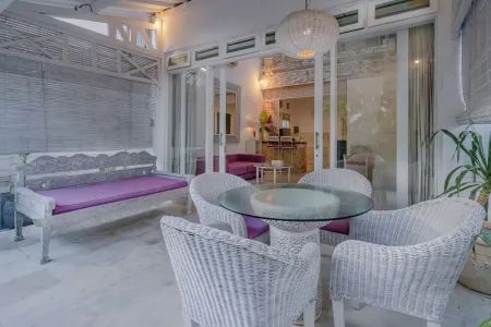 Elegant Balinese outdoor lounge with purple-cushioned daybed, glass-top wicker table and chairs, surrounded by potted plants and sliding glass doors. - Bali Villas
