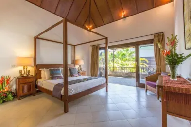Spacious tropical bedroom with wooden four-poster canopy bed, open sliding doors to balcony, and lush garden view.